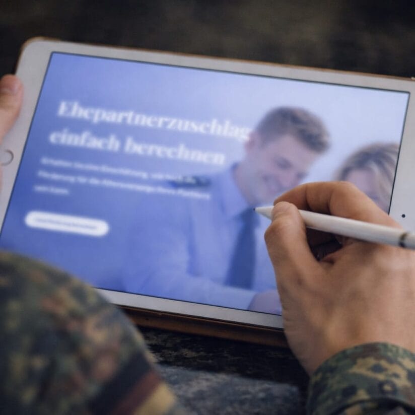 Person, die ein Tablet mit einem Stift in der Hand hält und eine deutsche Website zur Berechnung des Ehegattenzuschusses anzeigt, mit unscharfen Personen im Hintergrund.