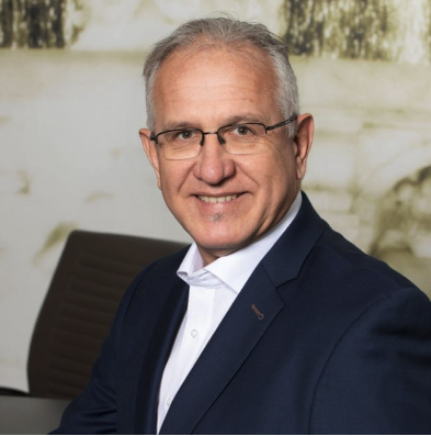 Ein Mann mit kurzen grauen Haaren und Brille, der einen marineblauen Anzug und ein weißes Hemd trägt, lächelt, während er in einem Bürostuhl sitzt.