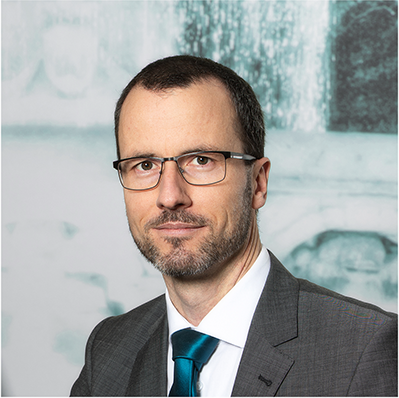 Ein Mann mit kurzem dunklem Haar, Brille und gestutztem Bart, der einen grauen Anzug, ein weißes Hemd und eine blaugrüne Krawatte trägt, posiert vor einem unscharfen Hintergrund.